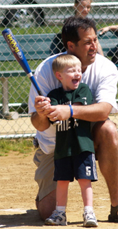 一张照片显示一个小男孩站在本垒棒球场举行铝棒和笑。孩子的父亲跪在一个膝盖身后,蝙蝠在同一立场的支持。